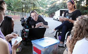 Image Description: Man in a wheelchair sitting around a small table with 4 other people holding playing card and playing a game. 